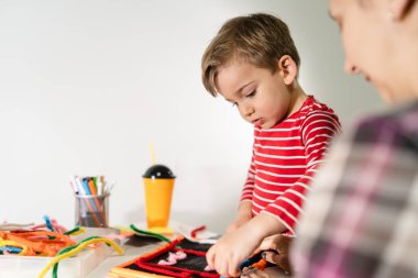 Küçük beyaz bir çocuğun omuzlarının üzerinden annesinin yanında duran ve evde masanın başında oynayan küçük bir çocuk.