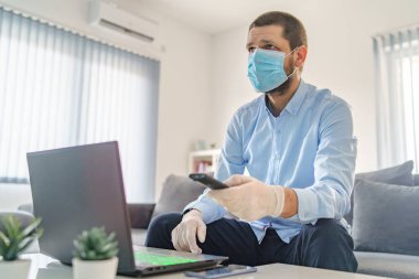 Koruyucu maske takan yetişkin bir beyaz adam elinde uzaktan kumandayla dizüstü bilgisayarın başında oturmuş maske takarak yayılan virüs salgını haberlerine bakıyor.