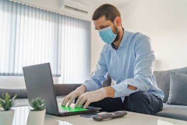 Ellerine eldiven takmış ve maske takmış genç bir beyaz adam evde oturmuş laptopun başında çalışırken salgın hastalığın karantina sağlığına yayılmasını önlüyor.