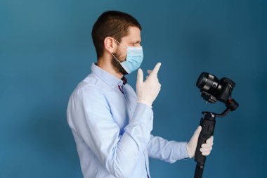 Kafkasyalı yetişkin blog yazarı, elinde kamerayla mavi duvarın önünde duruyor ve koruyucu maske takarak yayılan salgın virüsünün videosunu çekiyor.
