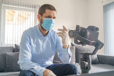 Kafkasyalı yetişkin blogcu gündüzleri evde oturmuş, karantinayı önlemek için koruyucu maske eldivenleriyle yayılan salgın virüsle ilgili video hazırlıyor.
