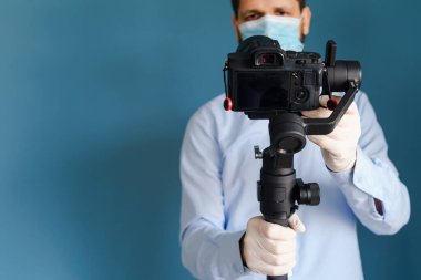 Kafkasyalı yetişkin blog yazarı, elinde kamerayla mavi duvarın önünde duruyor ve koruyucu maske takarak yayılan salgın virüsünün videosunu çekiyor.