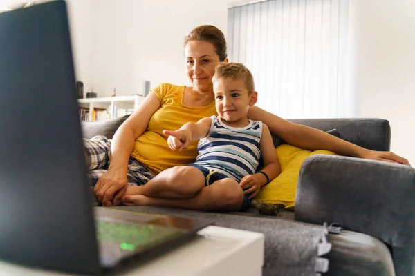 Small caucasian boy pointing finger to the screen shoving his mother while watching online content on laptop - Little child son sitting by his mom at home in front of the computer - childhood concept