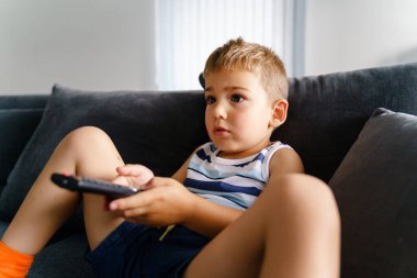 Küçük çocuk elinde uzaktan kumandayla kanepede oturuyor - küçük çocuk televizyon izliyor ve değiştiriyor - çocuk yaz günü evde dinleniyor - çocukluk aktivitesi konsepti