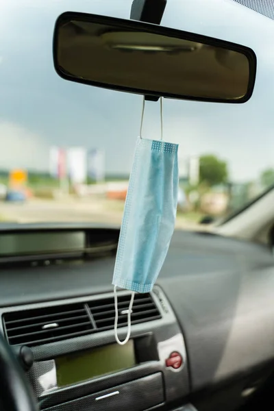 Medical protective face mask in car on the rearview mirror in day - Covid-19 pandemic protection on rear view in the vehicle - coronavirus epidemic new normal travel concept