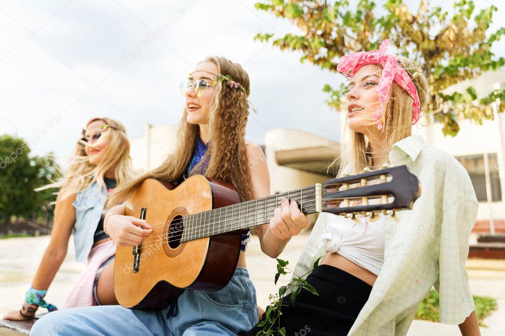 Young beautiful caucasian women playing guitar in summer day - three Pretty females friends or sisters in the city having fun singing - hippie style real people happiness and joy concept