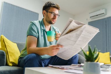 Yetişkin Kafkasyalı bir adam işten bir bardak kahve ya da çay içerken evde oturup gazete okuyor. Gerçek bir eğlence aktivitesi konsepti.