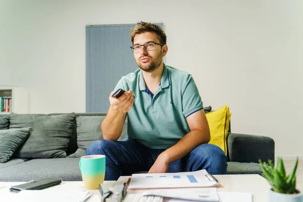 Front view on adult caucasian man sitting on the sofa bed at home using remote control - One male person changing tv channel while sit at home with chart documents on table - real people domestic life