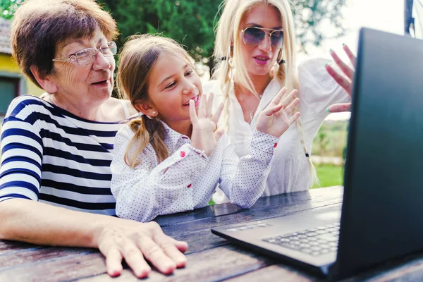 Female family grandmother mother daughter caucasian women and girl child using laptop in backyard to make a video call waving in summer day - Bonding and communication real people concept front view