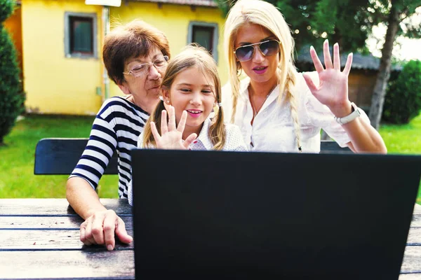 Female family grandmother mother daughter caucasian women and girl child using laptop in backyard to make a video call waving in summer day - Bonding and communication real people concept front view