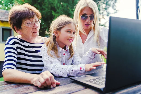 Female family grandmother mother daughter caucasian women and girl child using laptop in backyard to make a video call in summer day - Bonding and communication real people concept front view