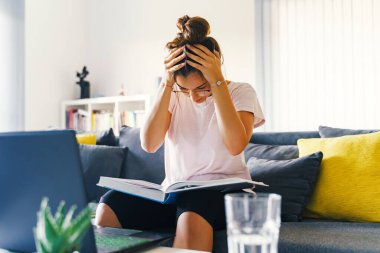 Kafkasyalı genç bir kadının evde ders çalışırken kafasını tutması, kanepede kitap okuması, yaz günü okul veya akademi sınavı için sorun yaşaması, eğitim konsepti.