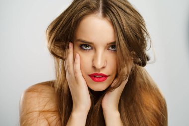 Güzel beyaz kız portresi kırmızı rujlu, esmer, doğal cilt ve makyajlı, çenesinde el ele tutuşmuş stüdyodaki kameraya bakıyor.