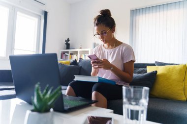 Kucağında kitapla evdeki kanepede oturan genç kız cep telefonu kullanıyor - akıllı telefonla internette gezinen kız öğrenci - eğitim ve eğlence konsepti