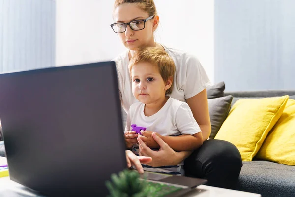 Single mother hold her little boy while using laptop at home - Caucasian woman with son watching video on notebook computer or making video call front view