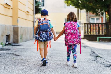 Küçük beyaz erkek ve kız kardeşlerin, kız ve erkek kardeşlerin ya da küçük arkadaşlarının arka plan portresi. Birinci sınıf öğrencileri güneşli bir günde sırt çantasıyla okuldan ayrılıyorlar.