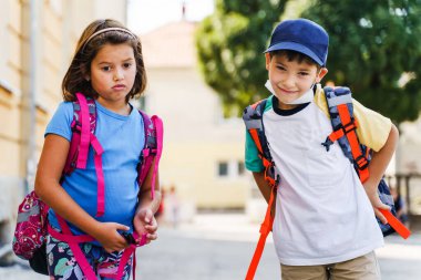 Küçük beyaz erkek ve kız kardeşlerin ön manzara portresi ya da küçük arkadaşlar okula giden birinci sınıf öğrencileri güneşli bir günde sırtlarında sırt çantalarıyla geziyorlar.