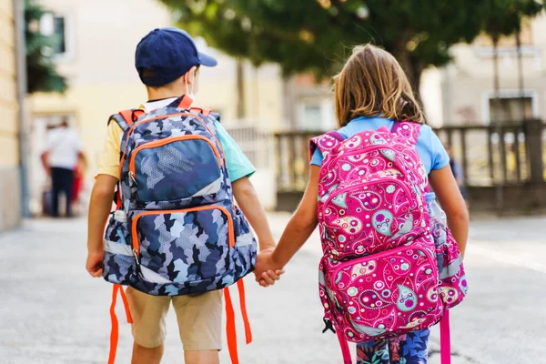 Güneşli bir günde sırt çantalı birinci sınıf öğrencilerini terk eden küçük beyaz erkek ve kız kardeşlerin ya da kız kardeşlerin tam portresi.