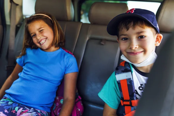 Small boy and girl brother and sister sitting on the back seat of the car n sunny day going to school looking to the camera happy making faces - Back to school education concept