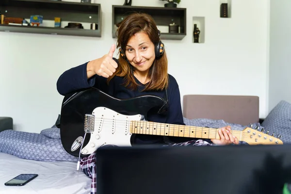 Caucasian woman with thumbs up having online class learning how to play guitar at home in her bedroom using laptop to develop new skill - Weekend activities real people leisure concept front view