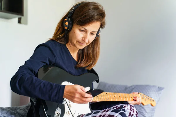 Adult caucasian woman sitting on bed with earphones on head using mobile phone to play music while holding guitar in her bedroom at home - real people leisure weekend activities concept
