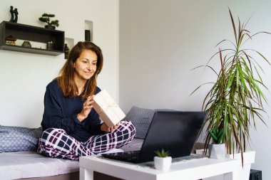Yetişkin beyaz bir kadının portresi evde pijamalarıyla yatarken dizüstü bilgisayarında hediye paketini gösteren bir video görüşmesi yapıyor. Sosyal mesafe kutlaması konsepti.