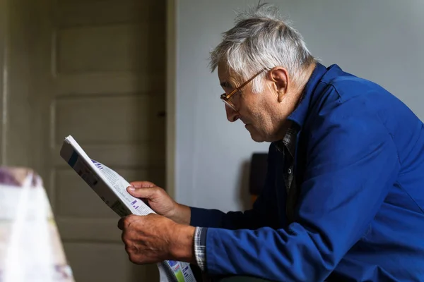 Side view of senior man reading newspapers at home in day - Old male pensioner peasant farmer sitting on bed at home reading press - retirement leisure and solitude concept