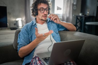 One man caucasian male freelancer work from home with headphones on his head on laptop computer customer support dispatcher while sit on sofa bed at home in shirt and pajamas frustrated confused tired clipart