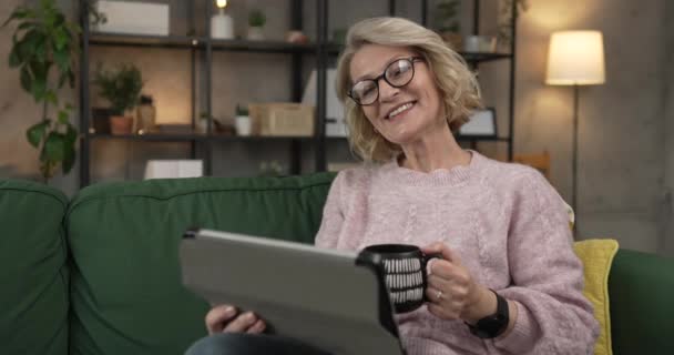 成熟したブロンドの白髪の女性の肖像画眼鏡は 家庭のインターネットでデジタルタブレットを使用するか オンラインビデオハッピー笑顔のスローモーションビデオを見る — ストック動画