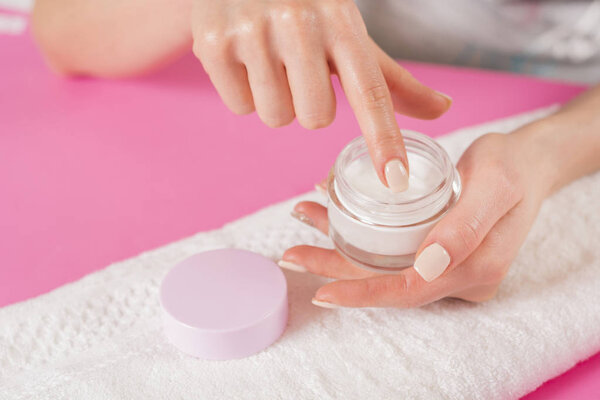 Woman finger touching open cream for hands on white towel and pink desk. Manicure and Hand beauty concept. Close up, selective focus