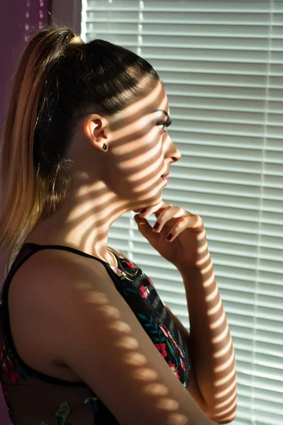 Beauty worrying woman in morning looking trough window and thinking. Vertical shadow of Venetian blinds falls on girl face