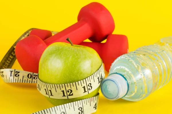 Green apple with measuring tape, red dumbbell and fresh water bottle with drops on yellow background. Healthy food and drink concept for sport and diet.