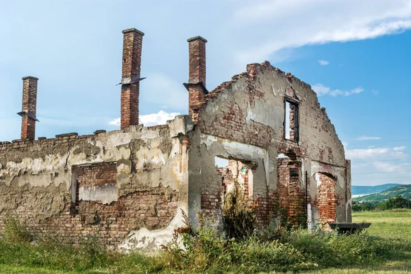 Abandoned destroyed old brick house without roof and with chimneys, broken windows, window frames, door and bricks surrounded with grass on cloudy weather day