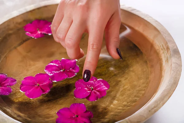 Young girl hand with purple varnish on nails touching flower in wooden bowl with water. Manicure and Spa beauty treatment concept. Selective focus, close up