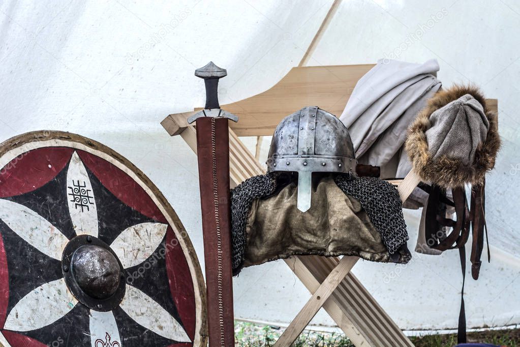Medieval knight equipment in old sleeping tent. Metal helmet, shield, sword and clothes on chair. Traditional knights weapons at middle age theme festival