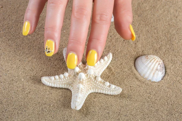 Female manicure with yellow nails polish on hand and starfish and shell on beach sand in background. Close up, selective focus