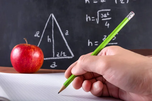 Erkek öğrenci el ile yeşil ahşap kalem not defterinde yazıyor. Kırmızı elma ve yazı tahtası arka planda bulanık. Sınıf ve eğitim kavramı. Yakın, seçici odak