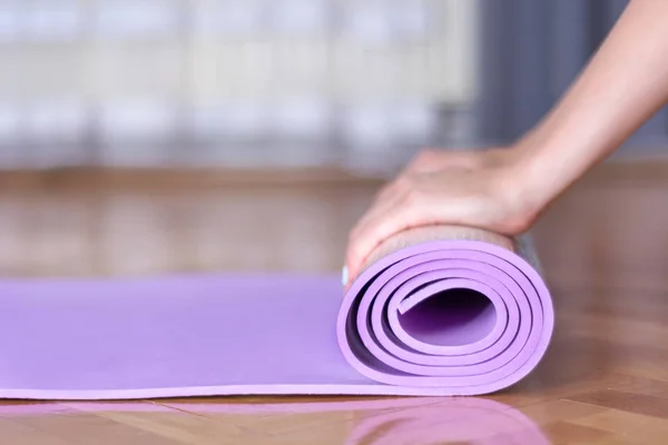 Young female hands roll yoga or fitness purple mat on parquet floor in the house. Sport and recreation concept. Close up, selective focus