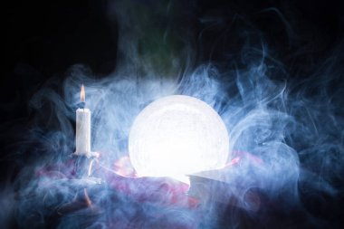 Fortune teller crystal ball smokes and lights on table with candle in candlestick and books. Halloween concept. Close up, selective focus clipart