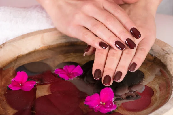 Young girl hands with brown manicure gel polish on fingers above water with violet flowers and black stones in wooden bowl in studio. Close up, selective focus