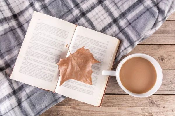 Open book with dry fallen leaf and cup of coffee on wooden background and retro blanket. Autumn concept image. Top view, selective focus