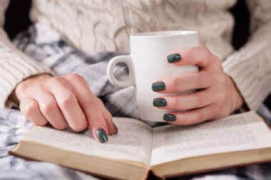 Kadın kitap ve holding fincan çay okuma. Kız yün kazak giyer ve retro bir battaniye ile örtülü. Kış ve soğuk hava kavramı. Yakın, seçici odak