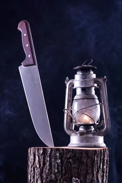 Big knife stabbed in wood log and old burning gas lantern. Smoky and foggy dark background. Horror and Halloween concept. Close up, selective focus