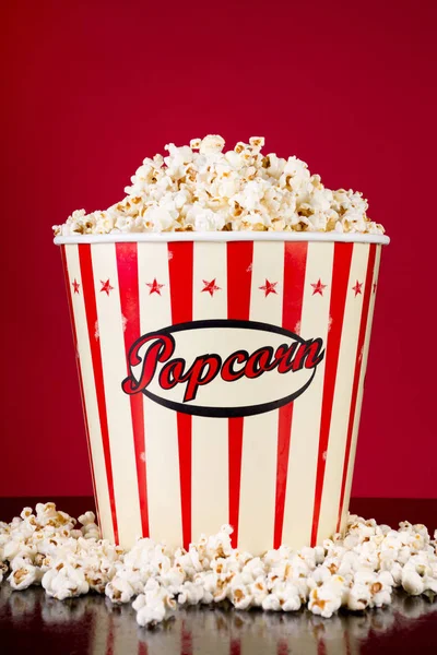 Retro full box with popcorn and spilled on black reflective desk and red background. Movie and cinema food concept. Close up