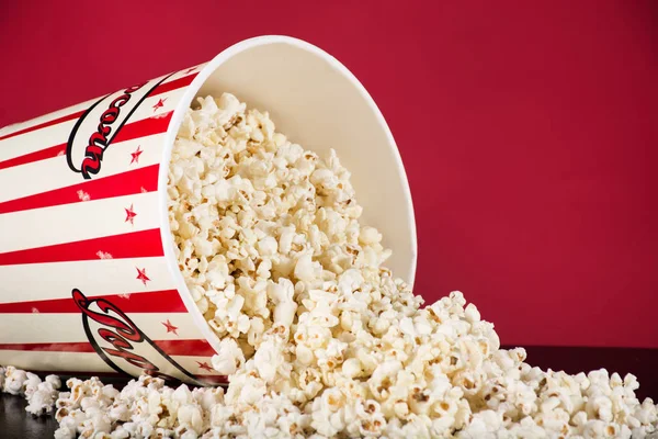 Spilled popcorn box on black desk and red background. Retro white and red box. Cinema and movie food concept. Close up, selective focus