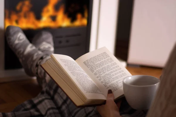 Woman reading book and hold cup of tea and warming feet in front of the fireplace at home in long winter night. Winter and cold weather concept. Close up, selective focus