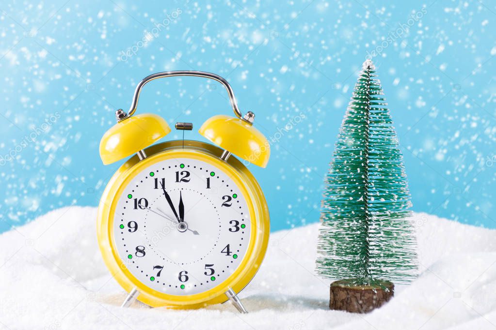 Retro clock and Christmas fir tree on snow and it's snowing winter day. Clock is in yellow color and snowflakes falling. Christmas and New Year holiday time concept