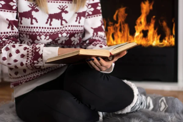 Girl sitting in front of the fireplace and reading book in long winter night at home. Girl warming and wears woolen socks and sweater. Winter and cold weather concept. Close up, selective focus