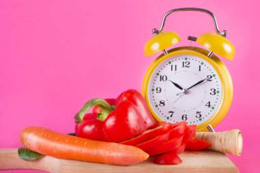 Taze sebze bir ahşap mutfak kurulu ve retro saat pembe arka plan izole. Kırmızı biber, havuç, yabani havuç ve tabak salataya. Chrono diyet ve beslenme kavramı. Yakın, seçici odak