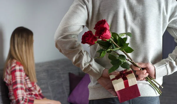 Mans hands hiding holding chic bouquet of red roses and gift with white ribbon behind back and woman with turned head awaits surprise in bed at home. Close up, selective focus